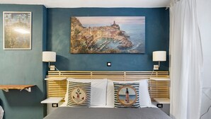 A bedroom with a bed, two bedside lamps, a headboard, a painting of a coastal town, and a small shelf. - Guest House 'Ca Du Grifun' with Wi-Fi and Air Conditioning (Vernazza)