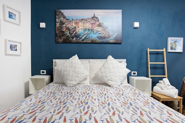 Un lit recouvert d’une courtepointe aux motifs de poissons colorés, avec des oreillers blancs, et un tableau représentant une ville côtière accroché au-dessus.