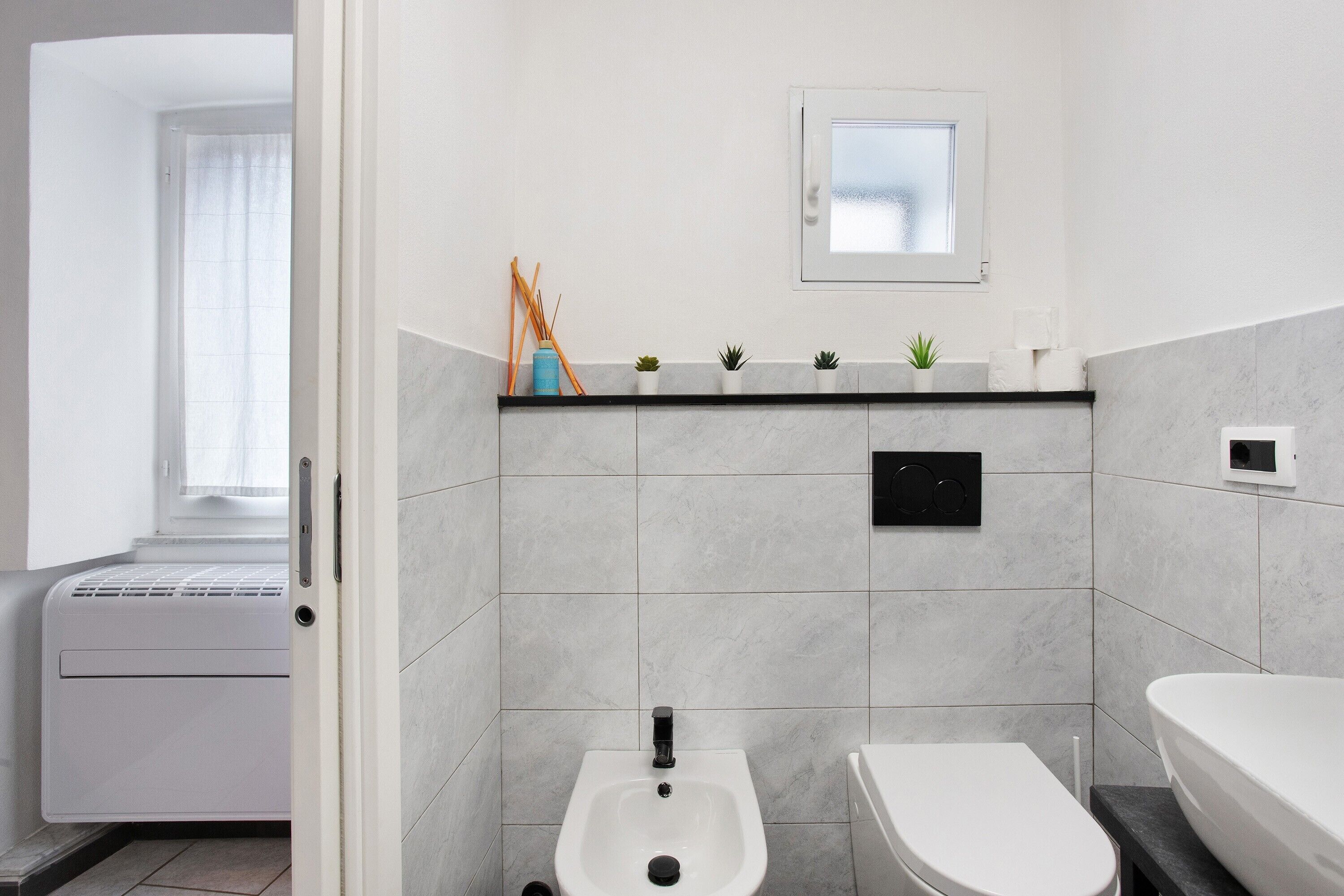 A modern bathroom with a bidet, toilet, and a shelf with plants and toiletries.