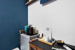 Quarto compacto com cozinha compacta, mini-geladeira, cafeteira, bandeja com xícaras e um controle remoto, e um código QR na parede.