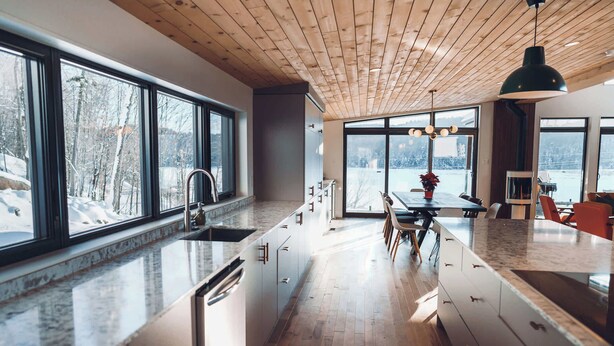 A modern kitchen with large windows, wooden ceiling, and a dining area with chairs.
