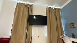 A room with a flat-screen TV mounted on the wall, beige curtains, a white desk with a chair, and a red couch. - Stylish 1BR Apartment Near Westbahnhof (Vienna)