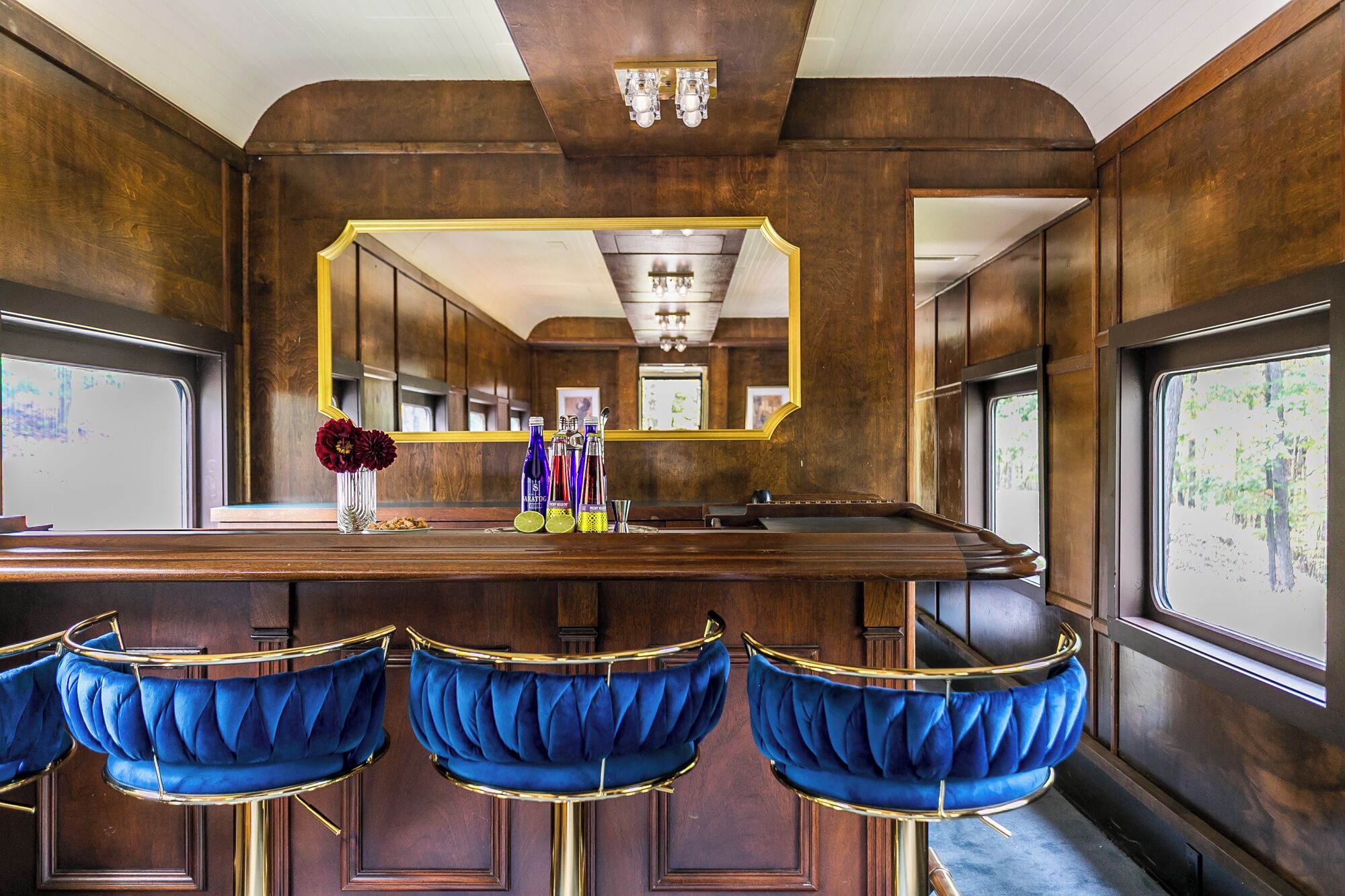 Un espace bar doté d’un comptoir en bois, de tabourets de bar en velours bleu et d’un grand miroir au-dessus du comptoir.