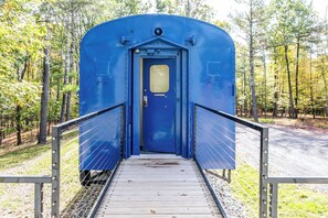 A blue portable restroom with a metal ramp leading up to the door, surrounded by trees. - Authentic 1940s Pullman Train Car Nestled in Bucolic Catskill Woods (Acra)