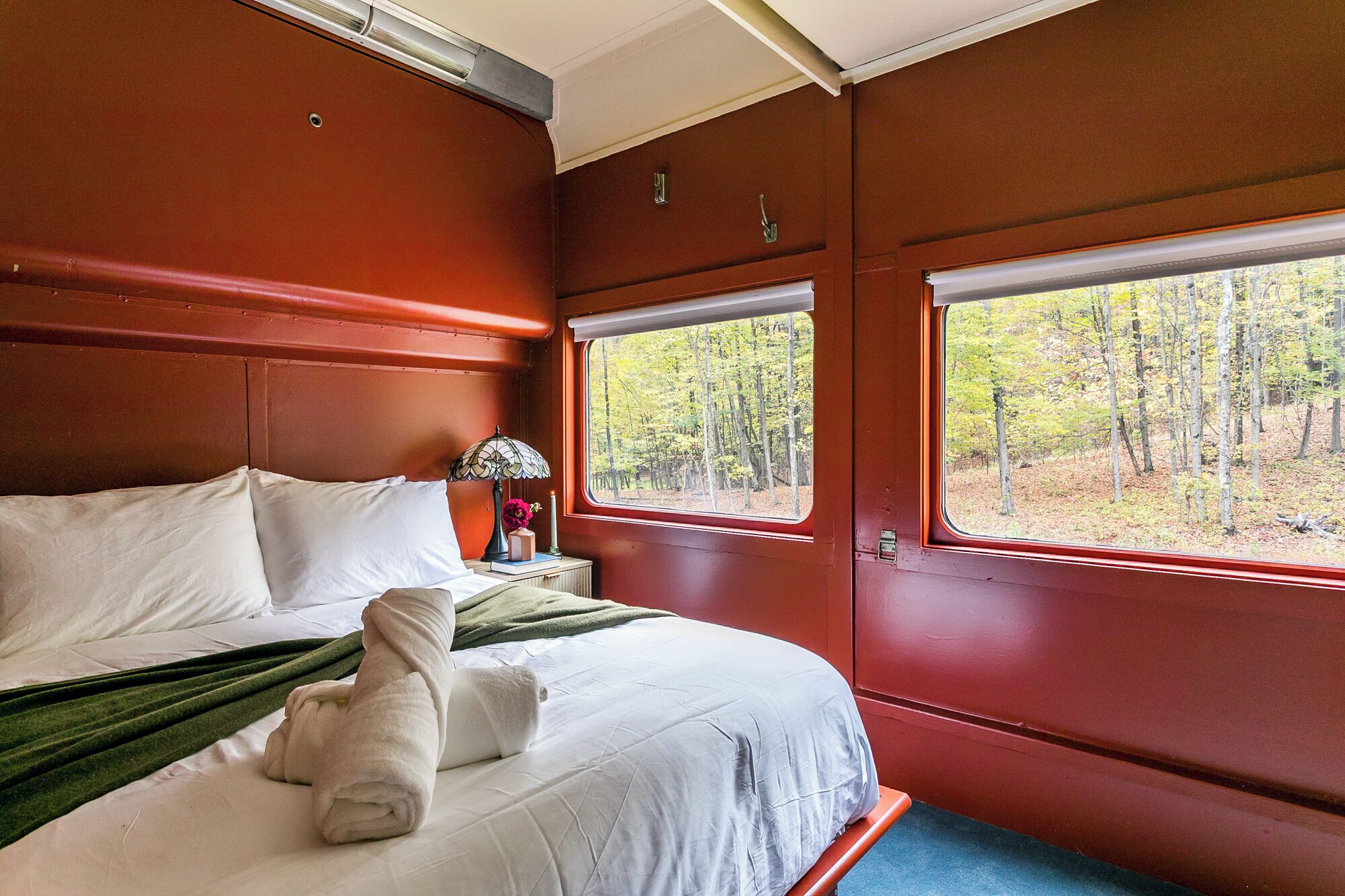 Une chambre à coucher avec un lit, une table de chevet, une lampe et une fenêtre donnant sur une forêt.