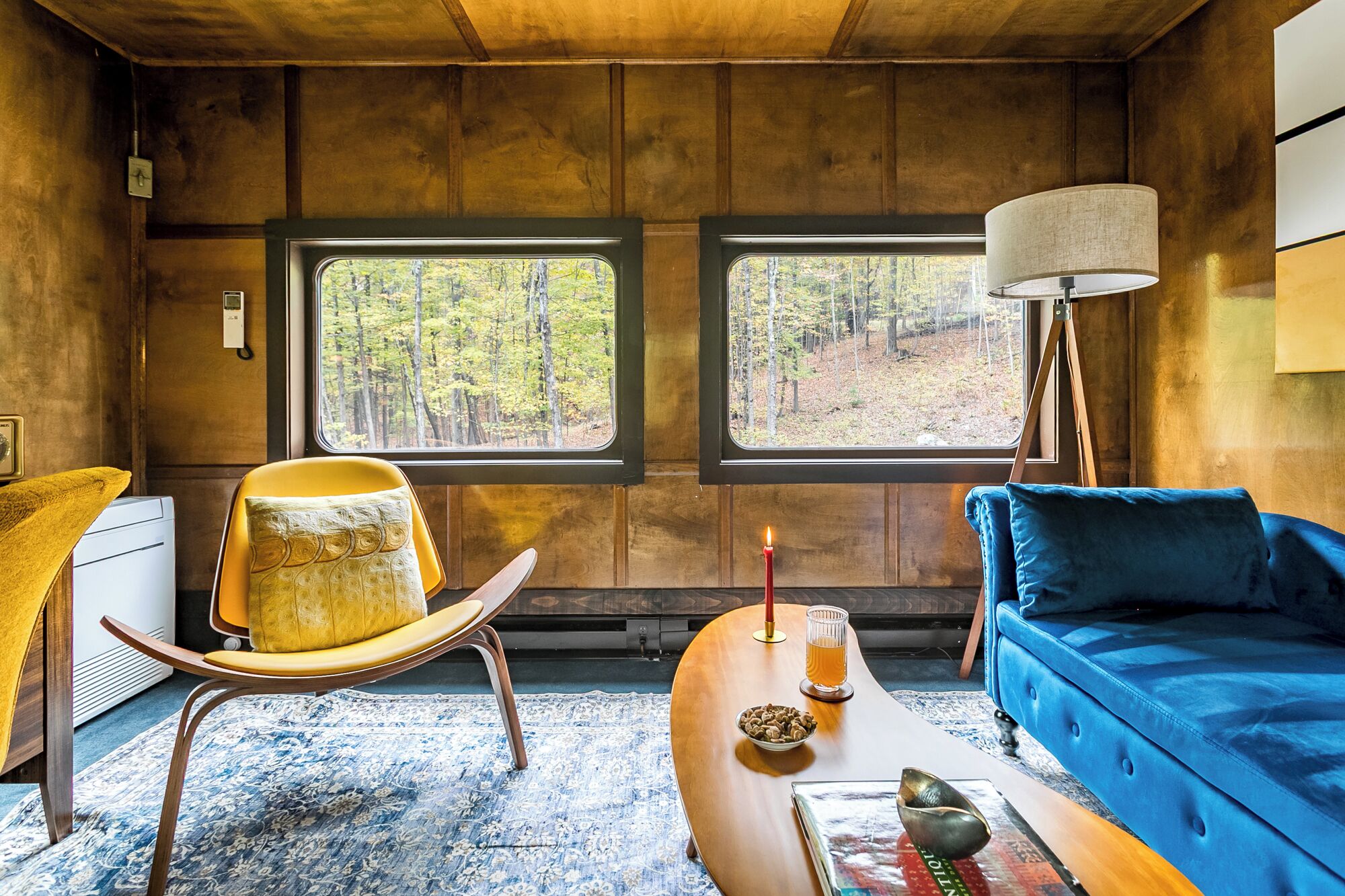 Une pièce chaleureuse avec un plafond en bois, deux fenêtres donnant sur la forêt, un fauteuil jaune, un canapé bleu, une lampe et une petite table sur laquelle se trouvent une bougie et un bol de noix.