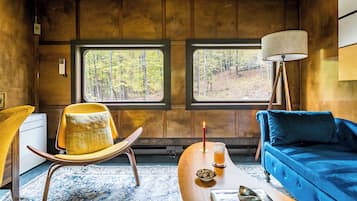 Une pièce chaleureuse avec un plafond en bois, deux fenêtres donnant sur la forêt, un fauteuil jaune, un canapé bleu, une lampe et une petite table sur laquelle se trouvent une bougie et un bol de noix.
