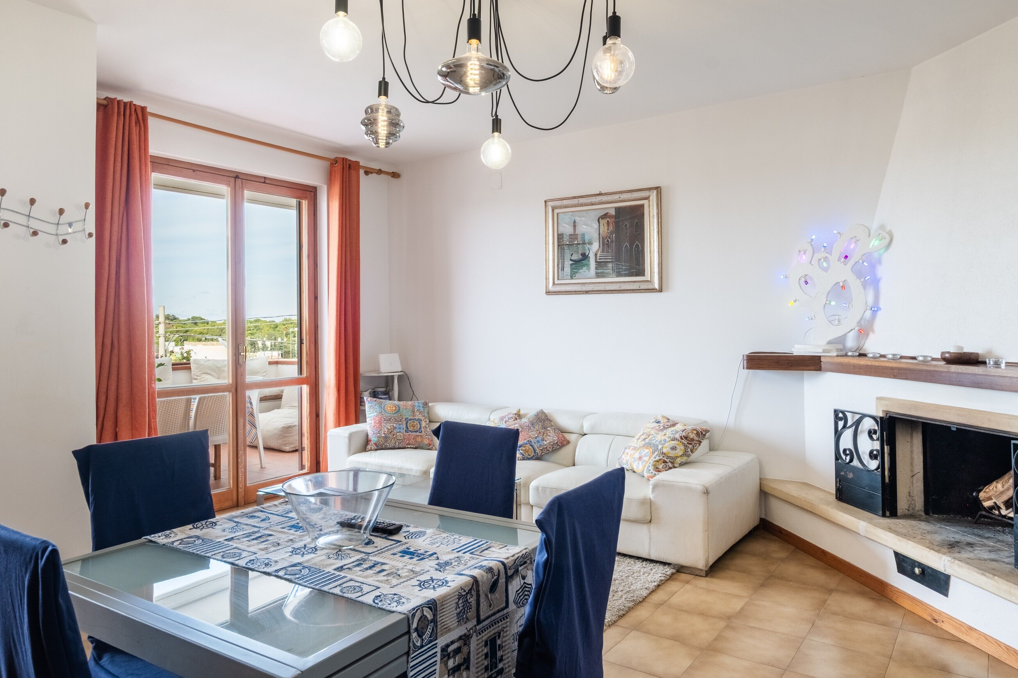 Una sala de estar con una mesa de vidrio, sillas azules, un sofá blanco, chimenea y un ventanal con cortinas.
