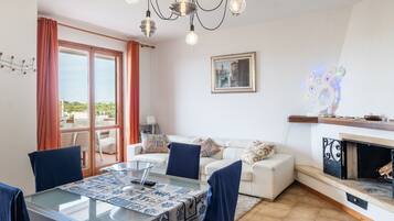 Una sala de estar con una mesa de vidrio, sillas azules, un sofá blanco, chimenea y un ventanal con cortinas.