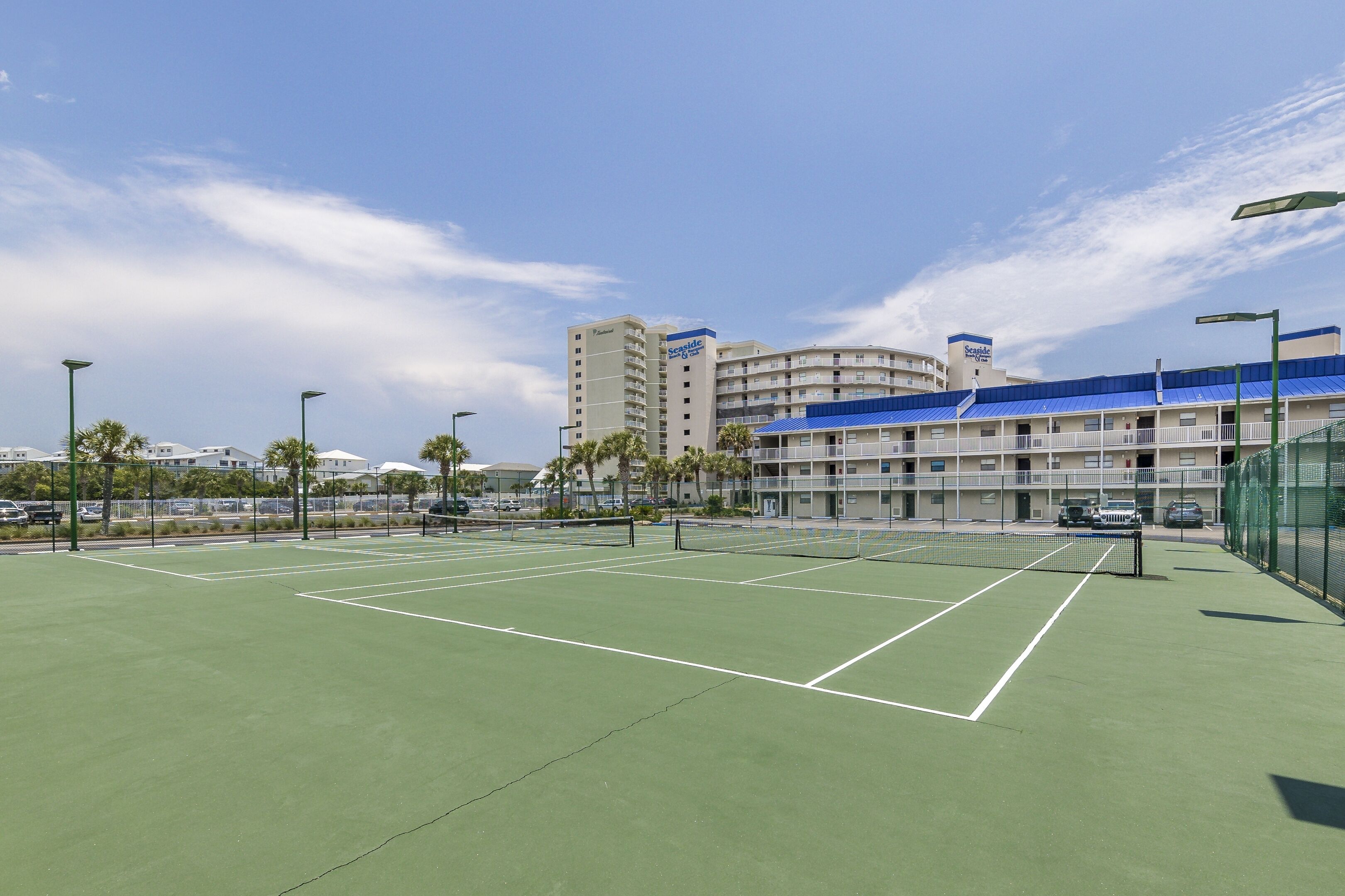 A tennis court with a net, surrounded by a fence, and buildings in the background.