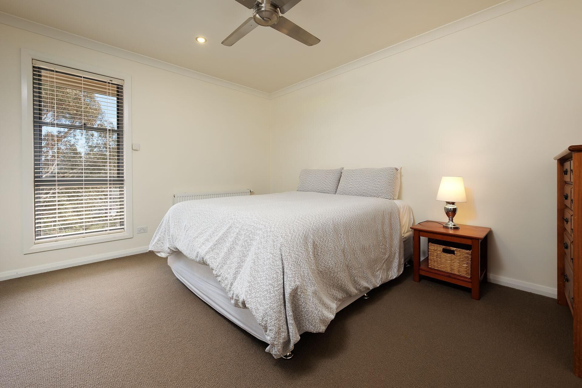 Quarto com cama, mesa de cabeceira com abajur, radiador e ventoinha no teto.