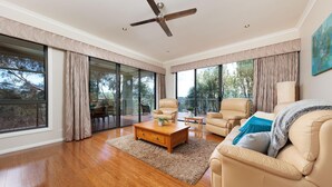 A spacious living room with a wooden floor, a ceiling fan, and large windows offering a view of the outdoors. - Peregrine Pool Paradise - Outstanding four bedroom family home with pool! (Wodonga)
