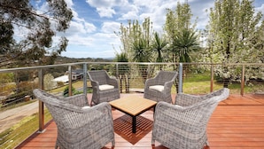 A balcony with wicker furniture and a wooden table, overlooking a garden with trees and a distant view of a house. - Peregrine Pool Paradise - Outstanding four bedroom family home with pool! (Wodonga)