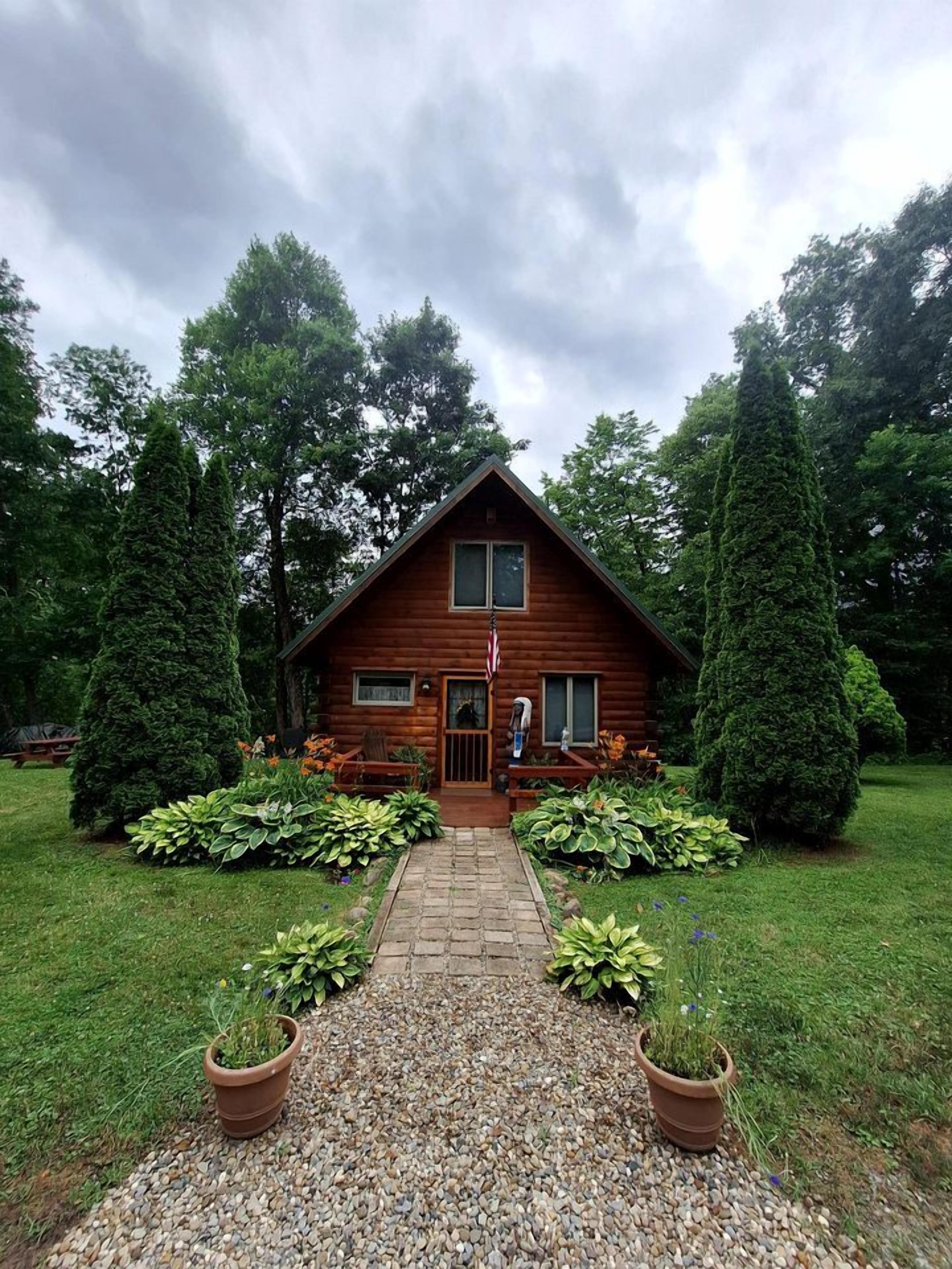 A wooden cabin with a central entrance, surrounded by well-maintained gardens and tall trees.