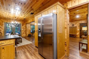 Innenansicht einer Holzhütte mit Küchenzeile, die einen Edelstahlkühlschrank, Holzschränke und einen Esstisch zeigt.