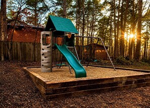 A playground with a slide and a climbing structure, surrounded by a wooden fence and trees. - Wonderful Cabin Perfect for Family Getaways in Broken Bow, Oklahoma (Broken Bow)