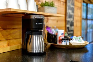 A Keurig coffee maker on a countertop with a wooden shelf above it, a plant, and a bowl with items on the counter.