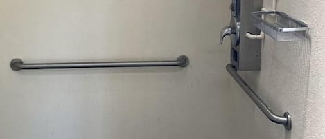 A bathroom with a grab bar, a shower control panel, and a metal shelf.