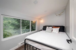 A bedroom with a large window, a bed with white bedding, a black headboard, and a bedside table.