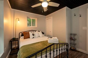 A bedroom with a bed, a nightstand, a bedside lamp, a window, a door, a ceiling fan, and a metal shelf unit. - Scenic Retreat: Riverfront Cabin with Large Deck and Fire Pit in Emlenton, Pennsylvania (Emlenton)