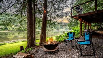 A lakeside outdoor seating area with a fire pit, two chairs, and a covered structure.