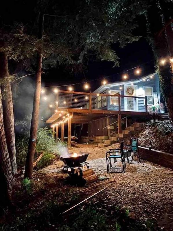 Une maison à deux étages avec une terrasse, entourée d’arbres et illuminée de guirlandes lumineuses.