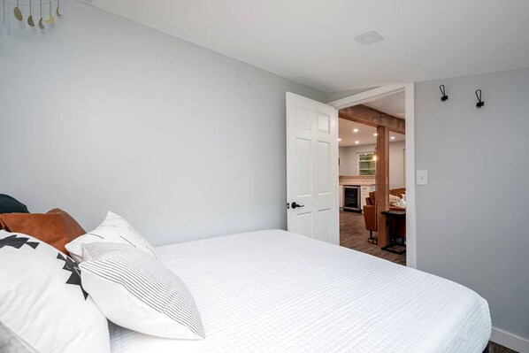 A bedroom with a bed, pillows, a door leading to a kitchen, and a light switch on the wall.