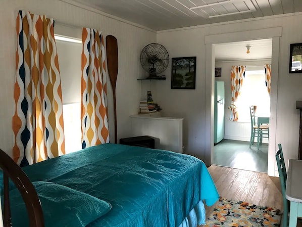 Une chambre à coucher avec un lit, des rideaux, un ventilateur, une étagère remplie de livres et une vue sur une autre pièce.