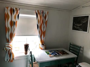 Une petite pièce avec une table et des chaises, une fenêtre avec des rideaux et un tableau accroché au mur.