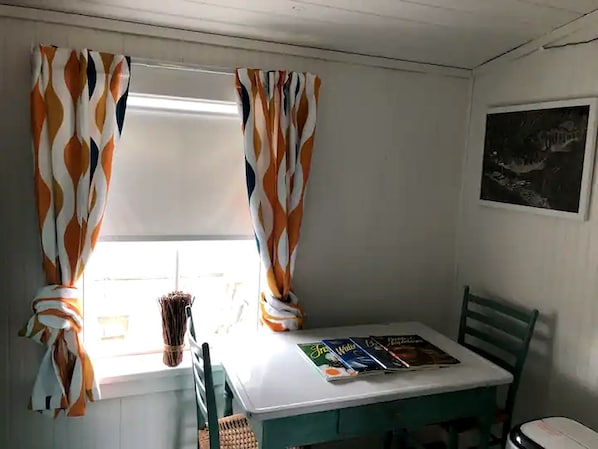 Una habitación pequeña con una mesa, sillas, un ventanal con cortinas y un cuadro en la pared.