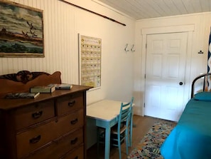 Une pièce comprenant une commode en bois, une petite table avec une chaise, un lit recouvert d’une couverture bleue, une porte et un mur orné d’un tableau encadré et d’un schéma.