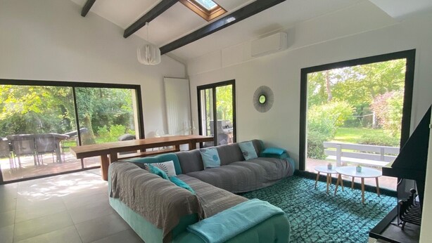 A modern living room with a sofa, a dining table, and a fireplace. There are skylights and large windows providing natural light.