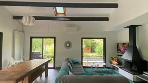 A modern living room with a wooden dining table, a sofa, and a TV. There are skylights and a fireplace.