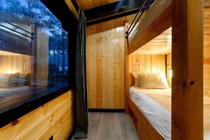 A cabin interior with a wooden floor, a bed with white bedding, a window with a view of trees, and a curtain.