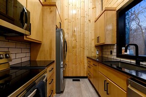 A compact kitchen with wooden cabinets, a stainless steel refrigerator, and a window with a view of trees.
