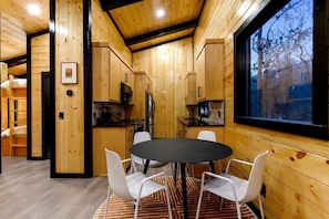 A wooden interior with a round table and chairs, a kitchen area, and a window with a view.