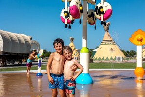 Two children standing in a water park with water features and a large tent in the background.