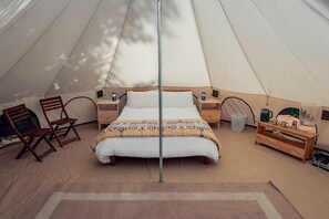 A tent with a bed, wooden chairs, a table with items, and a trash bin.