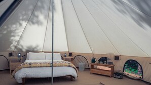 A tent with a bed, bedside tables, a lamp, and a window with a view of greenery.