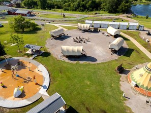 An aerial view of a recreational area with a circular water play area, covered storage units, and a small building with a distinctive roof. - Perfect Glamping Vacations in this Rustic Conestoga Covered Wagon Rental in Oklahoma (Moore)