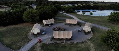 An outdoor seating area with multiple covered tables and chairs, surrounded by trees and a body of water.