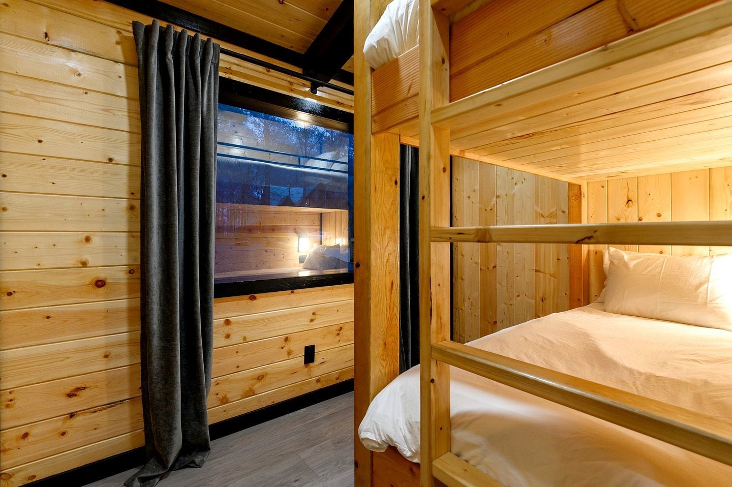 Una habitación de cabaña de madera con literas, una ventana con cortinas y vistas al exterior.
