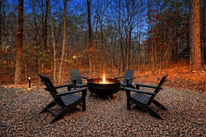 A fire pit with four chairs arranged around it, set on a gravel surface in a forested area.