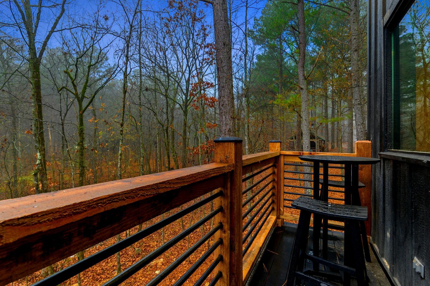 A wooden deck with a railing, a table, and chairs, overlooking a forested area.
