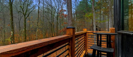 A wooden deck with a railing, a table, and chairs, overlooking a forested area.