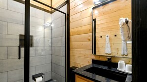 A modern bathroom with a glass shower enclosure, a wooden vanity with a black countertop, and a mirror reflecting a towel rack. - Garage Loft • King + Bunks • Budget Friendly near Beavers Bend National Park (Broken Bow)