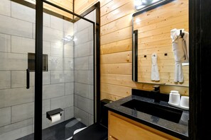 Ein modernes Badezimmer mit einer Duschkabine aus Glas, einem Holzwaschtisch mit schwarzer Arbeitsplatte und einem Spiegel, der einen Handtuchhalter reflektiert.