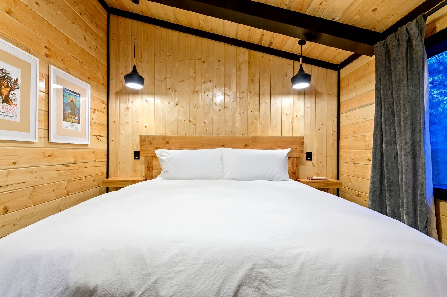 A wooden-paneled room with a bed, two pendant lights, and framed pictures on the wall.