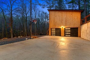 A modern wooden building with a basketball hoop, two garage doors, and a concrete driveway. - Garage Loft • King + Bunks • Budget Friendly near Beavers Bend National Park (Broken Bow)