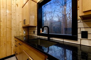 A kitchen with wooden cabinets, a black countertop, and a window with a view of trees. - Garage Loft • King + Bunks • Budget Friendly near Beavers Bend National Park (Broken Bow)